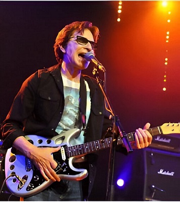 Russ Ballard con su guitarra personalizada con agujeros, cantando en un escenario